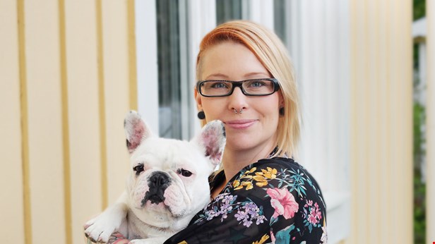 Anna står framför en gul vägg. Hon ler mot kameran och håller en vit hund i famnen..