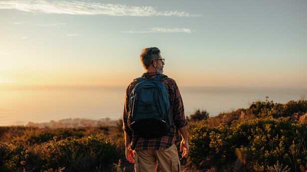 Man står i fjällnatur och vatten i horisonten. Han har ryggsäck på ryggen och står med ryggen vänd mot kameran. foto Istock